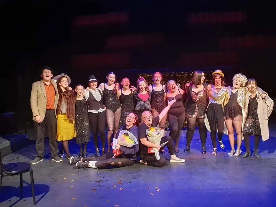 Group of performers on stage celebrating after a show, some holding flowers, smiling and posing for a photo.