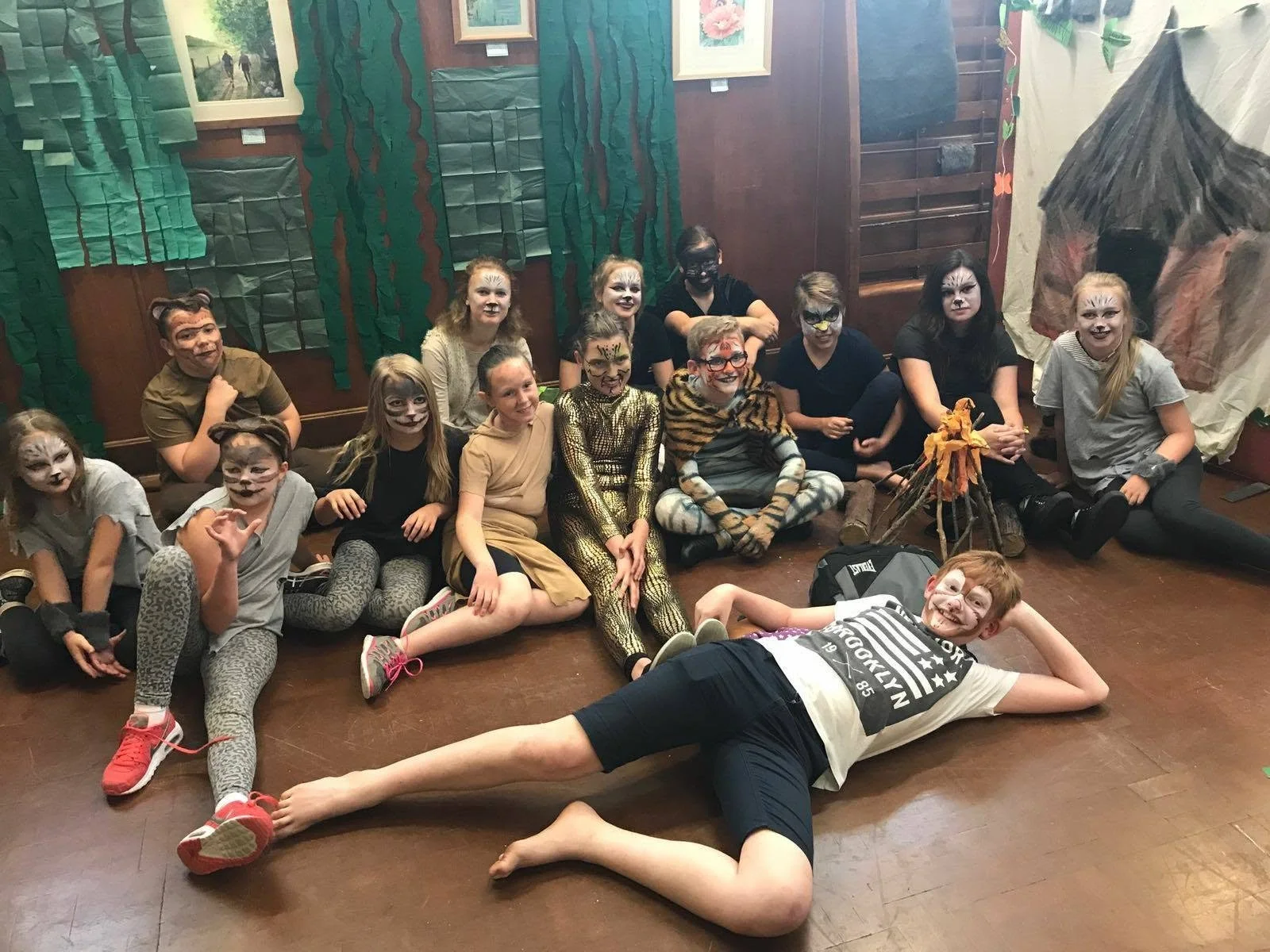 A group of children and teens in Halloween costumes with face paint, sitting and lying on the floor in a room decorated with green and brown handmade paper decorations and artwork.