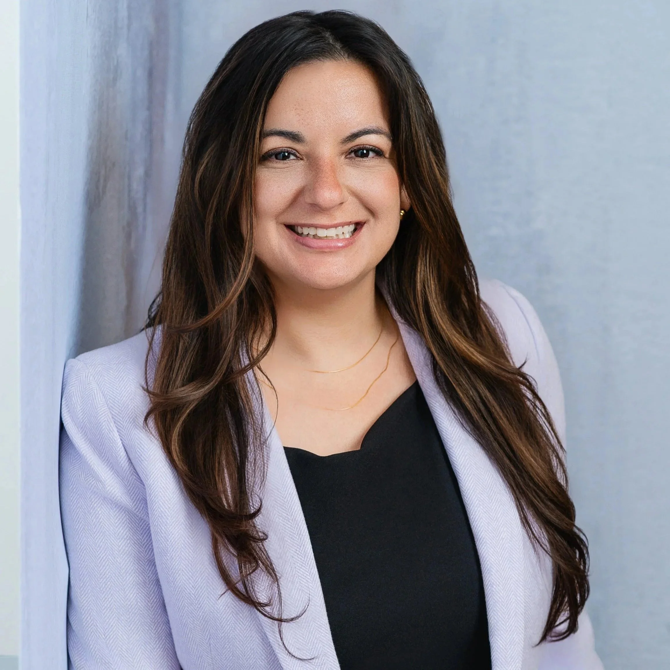 A woman with long, dark, wavy hair smiling, wearing a light purple blazer and a black top, standing against a neutral background.