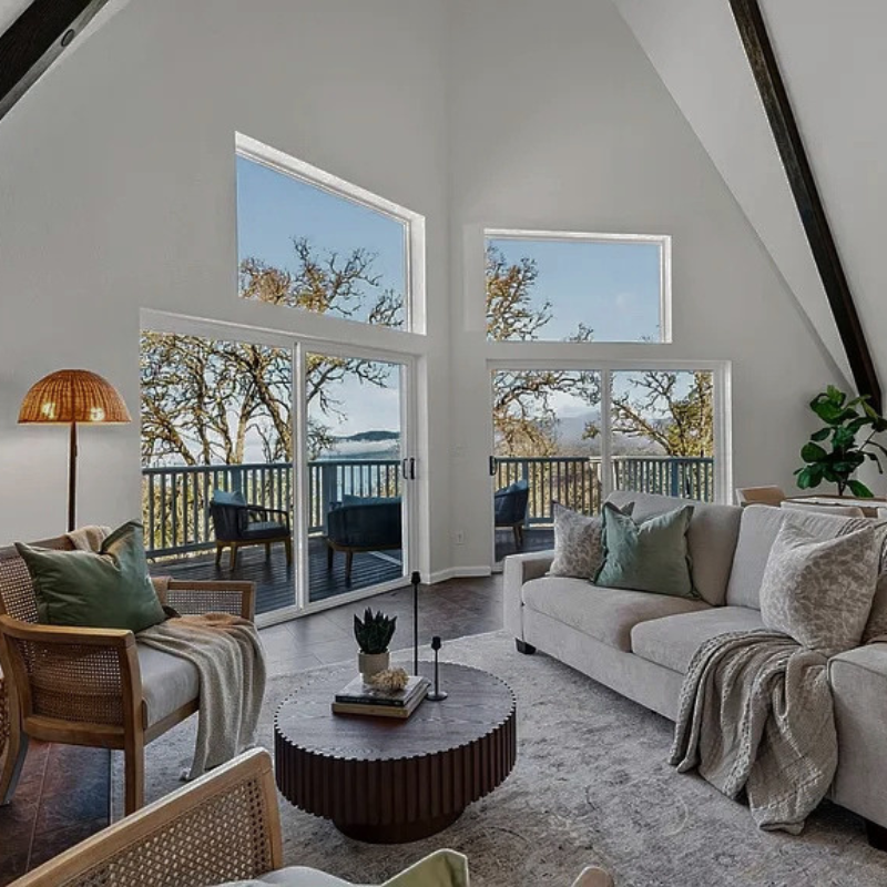 Living room with large windows and sliding glass doors overlooking a balcony and trees outside. Furniture includes a white sofa with pillows, a wooden armchair with a cushion and throw blanket, and a round coffee table with decorative items. There is a floor lamp and potted plant in the room.