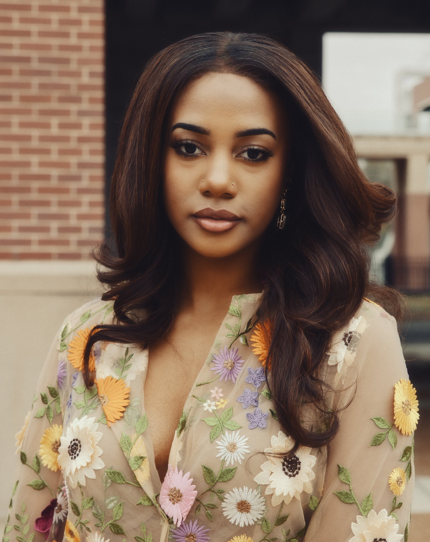 Close-up of a woman with long brown curled hair, wearing a sheer beige top with colorful embroidered flowers. She has dark makeup, hoop earrings, and a small nose piercing. The background shows a brick wall and a building.