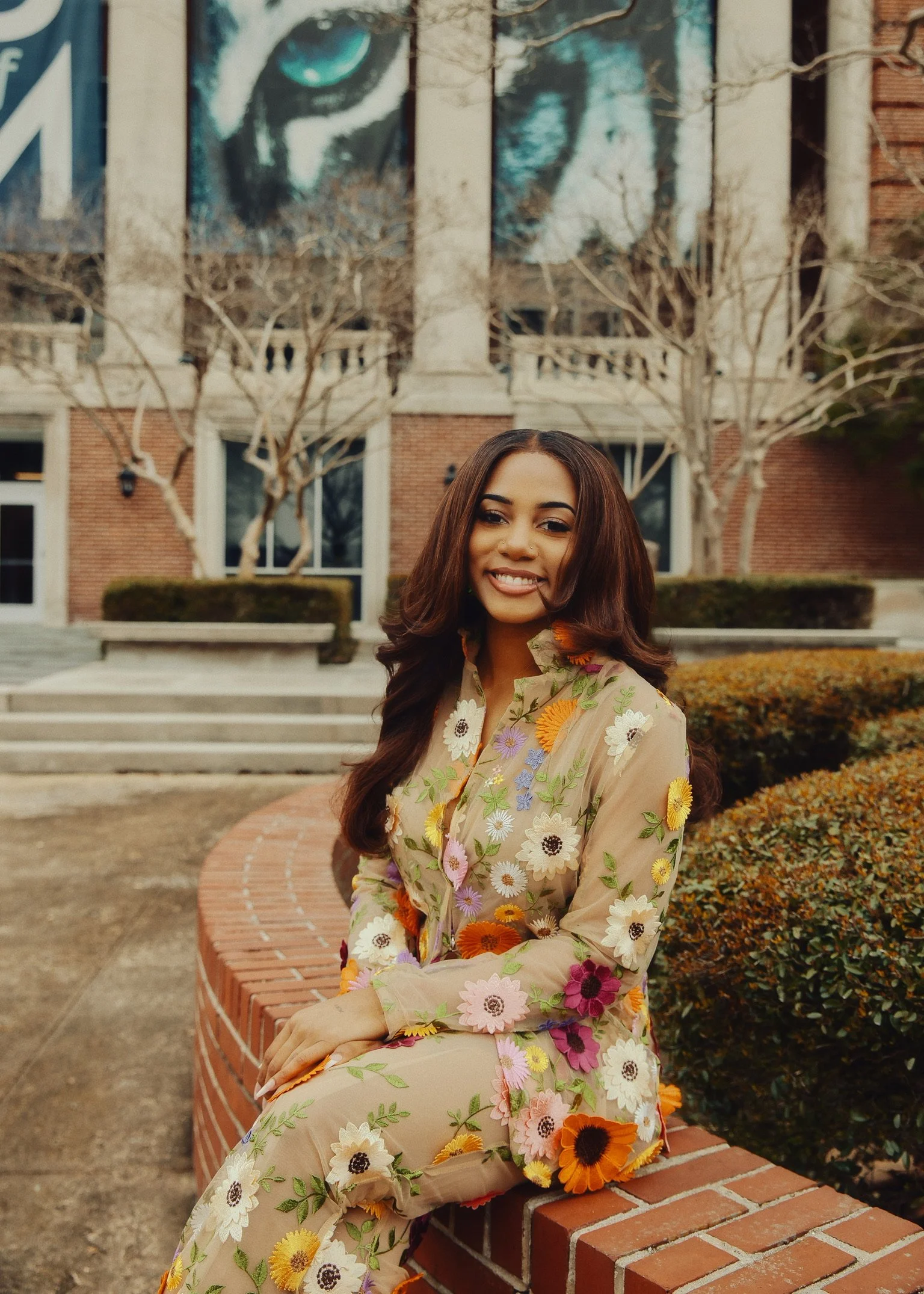A young woman with long brown hair sitting on a brick wall in front of a building with large windows and a mural. She is wearing a sheer beige dress decorated with colorful embroidered flowers and is smiling at the camera.