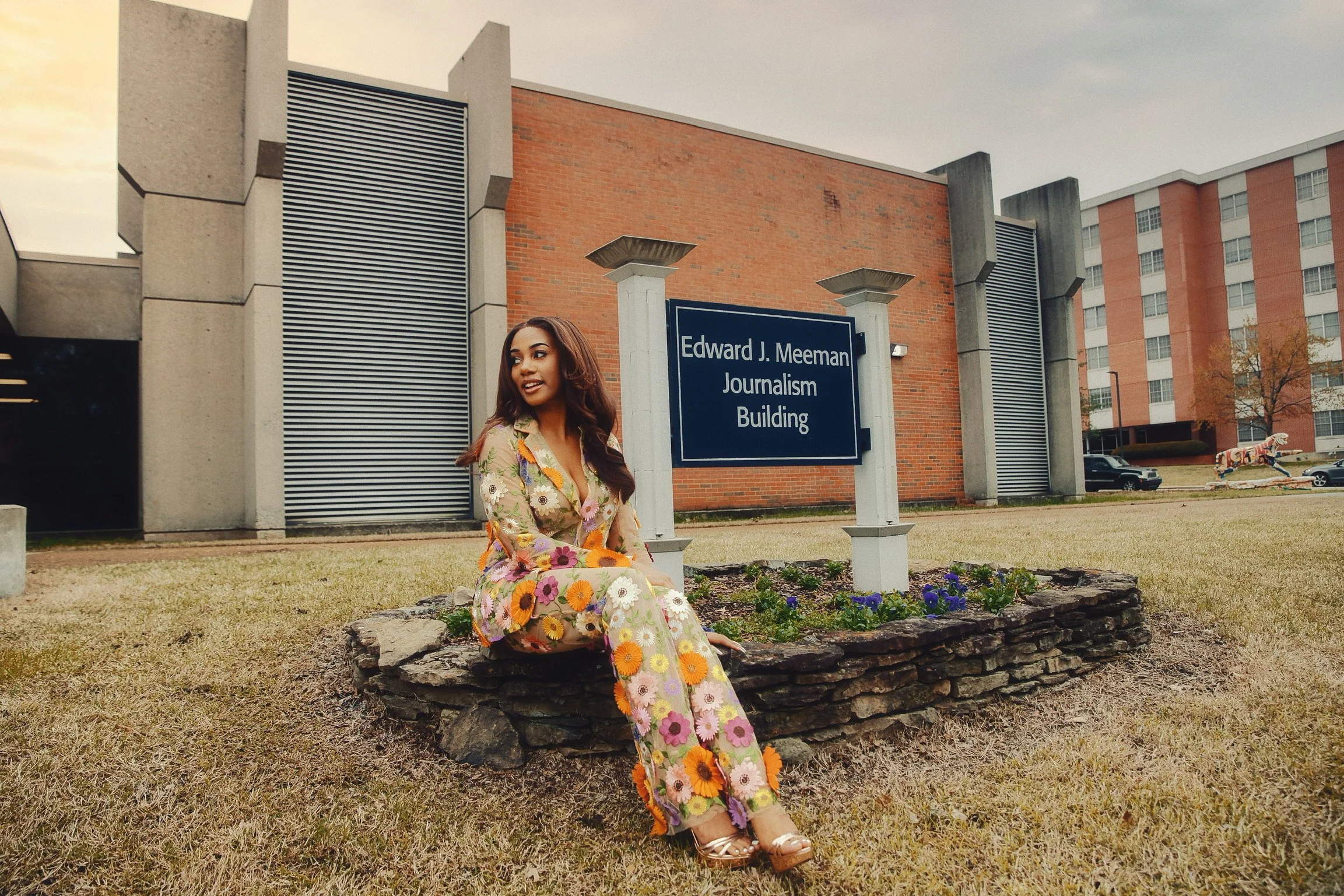 A woman in a floral dress is sitting on a stone border in front of a campus building with a sign that reads 'Edward J. Meeman Journalism Building.' She is outdoors on a grassy area with flowers and trees in the background.