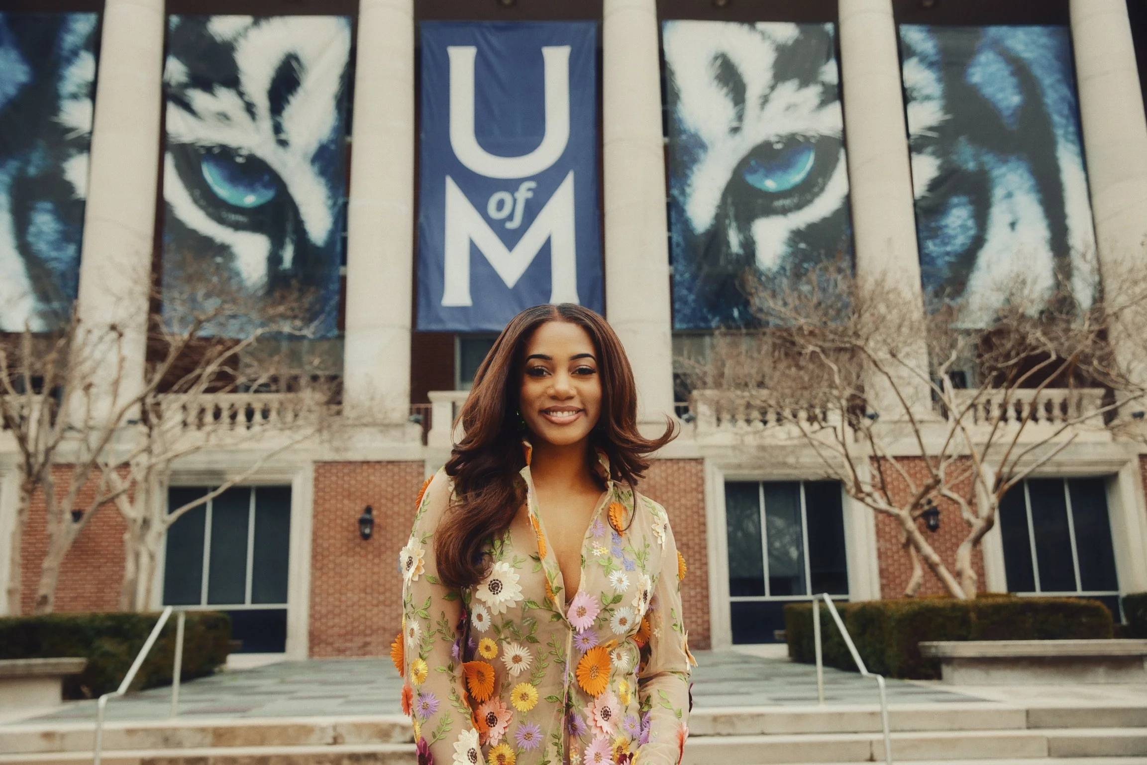 A young woman with long, dark hair and a floral dress standing in front of a building that has large banners with eyes and a sign that says "U of M". The scene includes leafless trees and steps leading to the entrance.