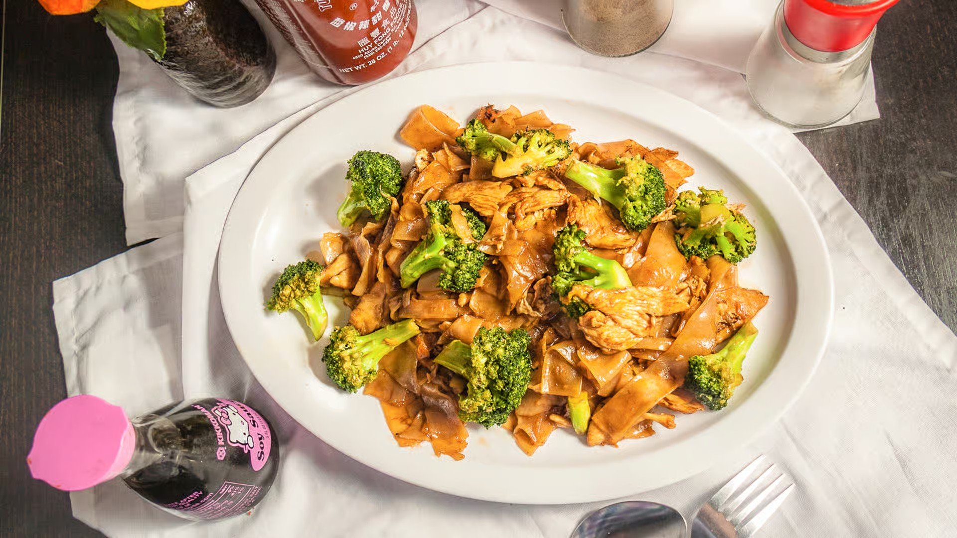 A plate of Asian-style stir-fried noodles with broccoli and chicken, on a dark table with condiments and drinks around it.