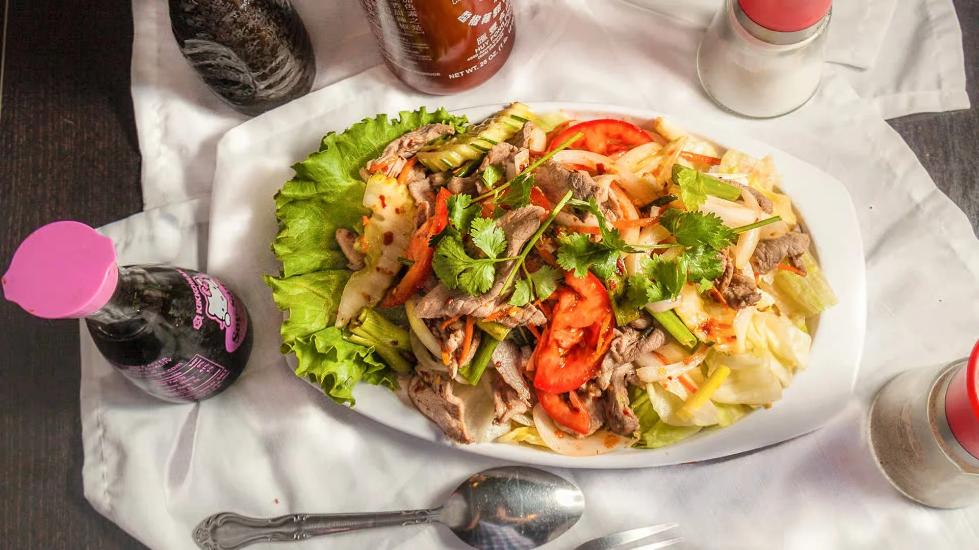 A plate of Asian-style noodle salad topped with sliced beef, chopped vegetables, herbs, and lettuce leaves. The plate is on white paper, accompanied by bottles of soda and condiments on a dark table.