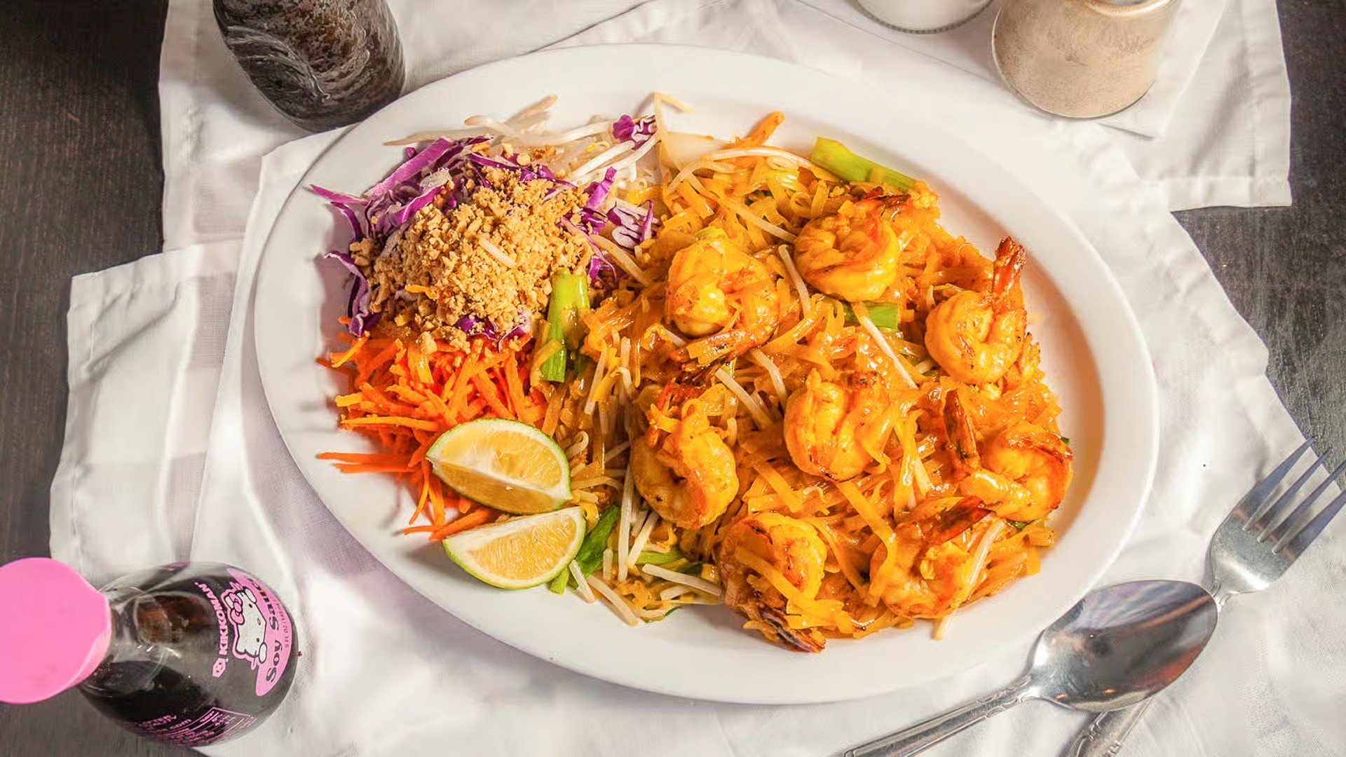 A plate of shrimp fried rice with lime wedges, shredded carrots, bean sprouts, shredded cabbage, and crushed peanuts, served on a white tablecloth with a fork, spoon, and drinks nearby.