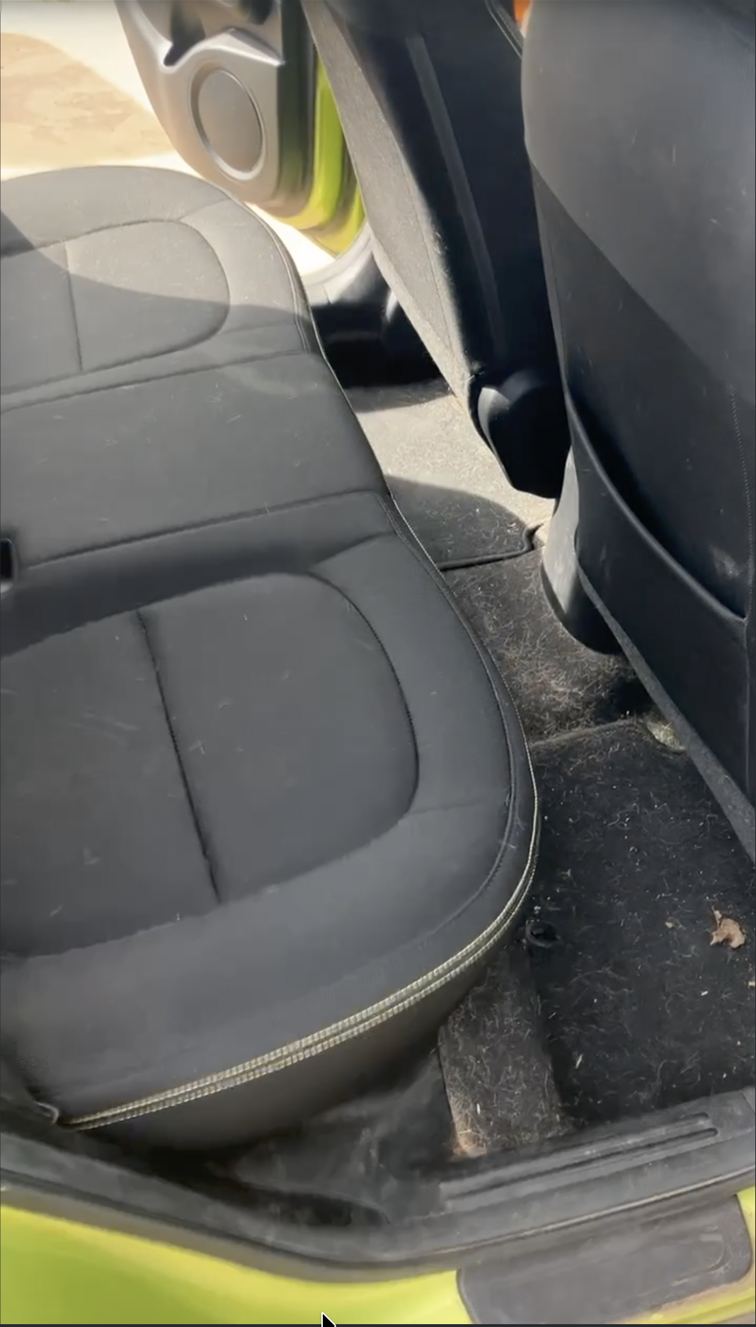 The backseat of a vehicle with black seat upholstery and a black floor, with some dirt and debris on the floor. The seat is slightly dusty with a textured design.