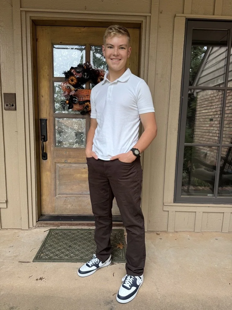 A young man standing on a porch in front of a wooden door with a wreath. He's wearing a white polo shirt, dark pants, and sneakers, with hands in pockets and smiling.