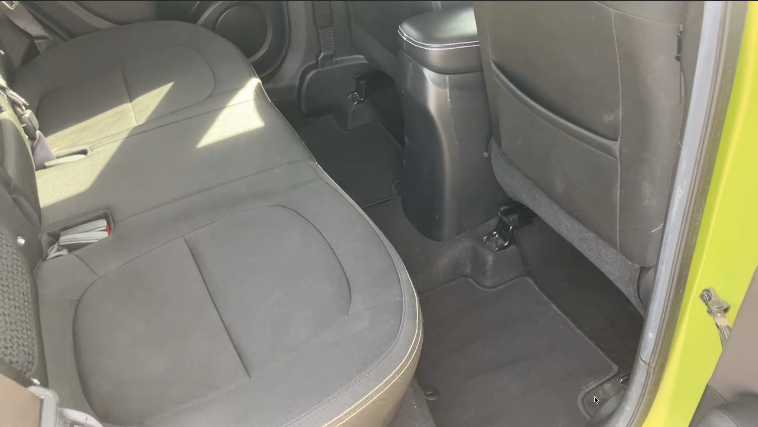 Interior of a vehicle showing the backseat area, with black fabric seats and a gray fabric back of the front seats. There are seat belts, and the floor is covered in black carpeting. The image also shows part of a yellow door frame on the right side.
