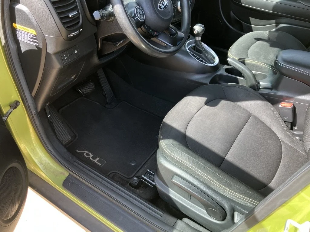 Interior view of a Kia Soul car showing the driver's seat, steering wheel, gear shift, brake and accelerator pedals, and part of the dashboard and door interior.