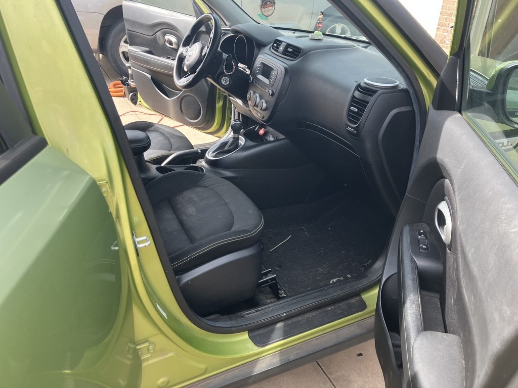 The interior of a green car showing the driver's side with a black steering wheel, dashboard, gear shift, and black fabric seat, with the door open revealing the door panel with window controls.
