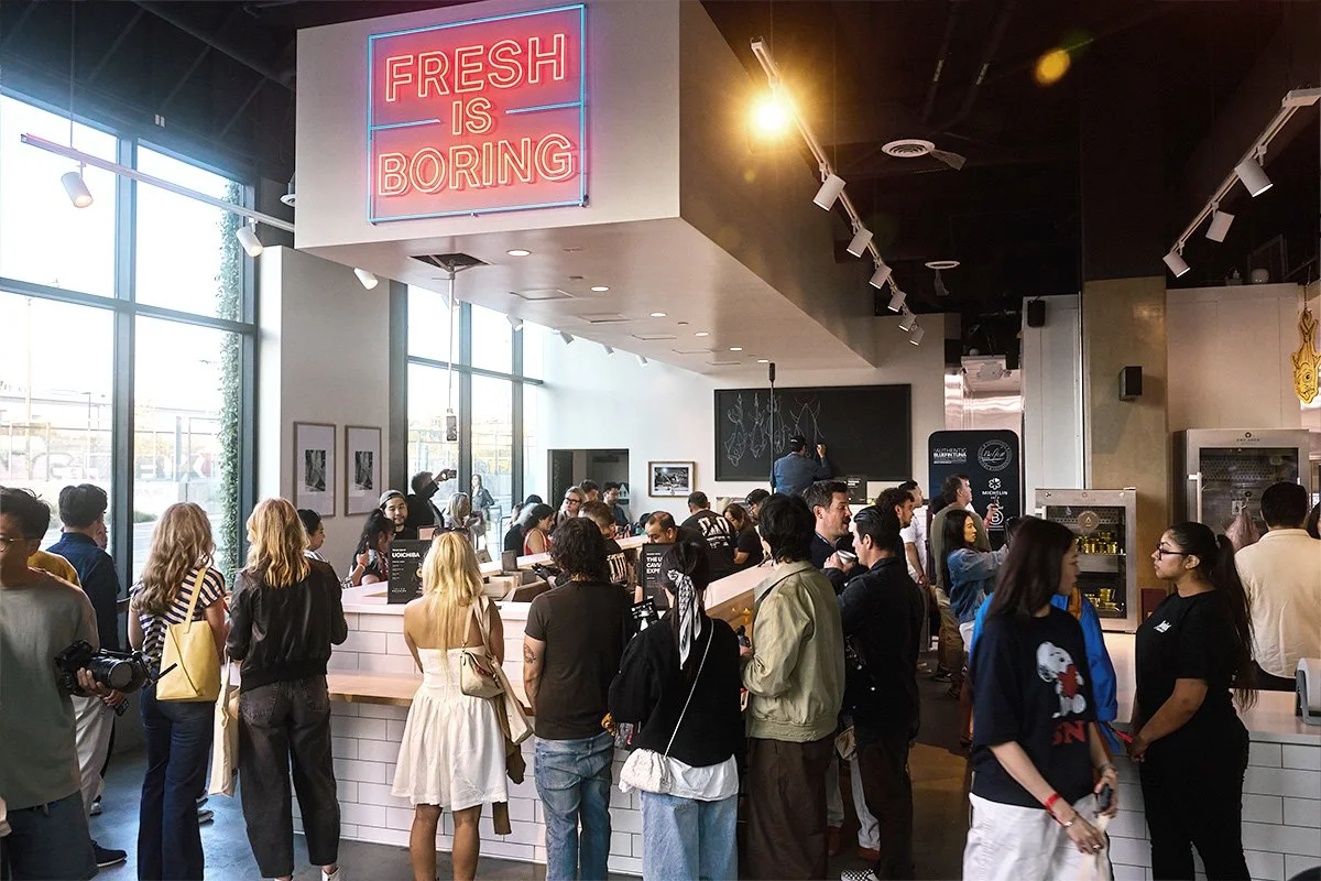 Crowd of people inside a modern clean handroll bar with large windows, a neon sign that reads 'FRESH IS BORING,' and a counter where customers are being served.