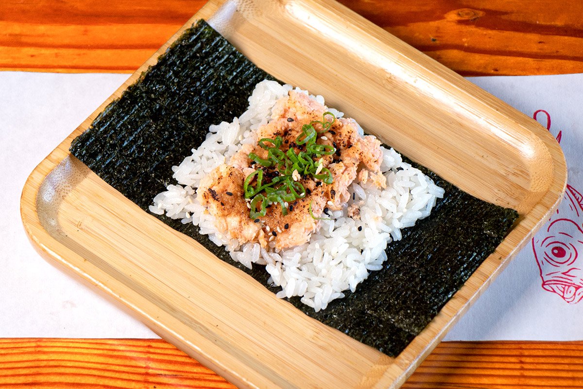 A bamboo tray showcasing an open handroll with roasted salmon, teriyaki, scallions and sesame on rice (shari) and seaweed (nori).
