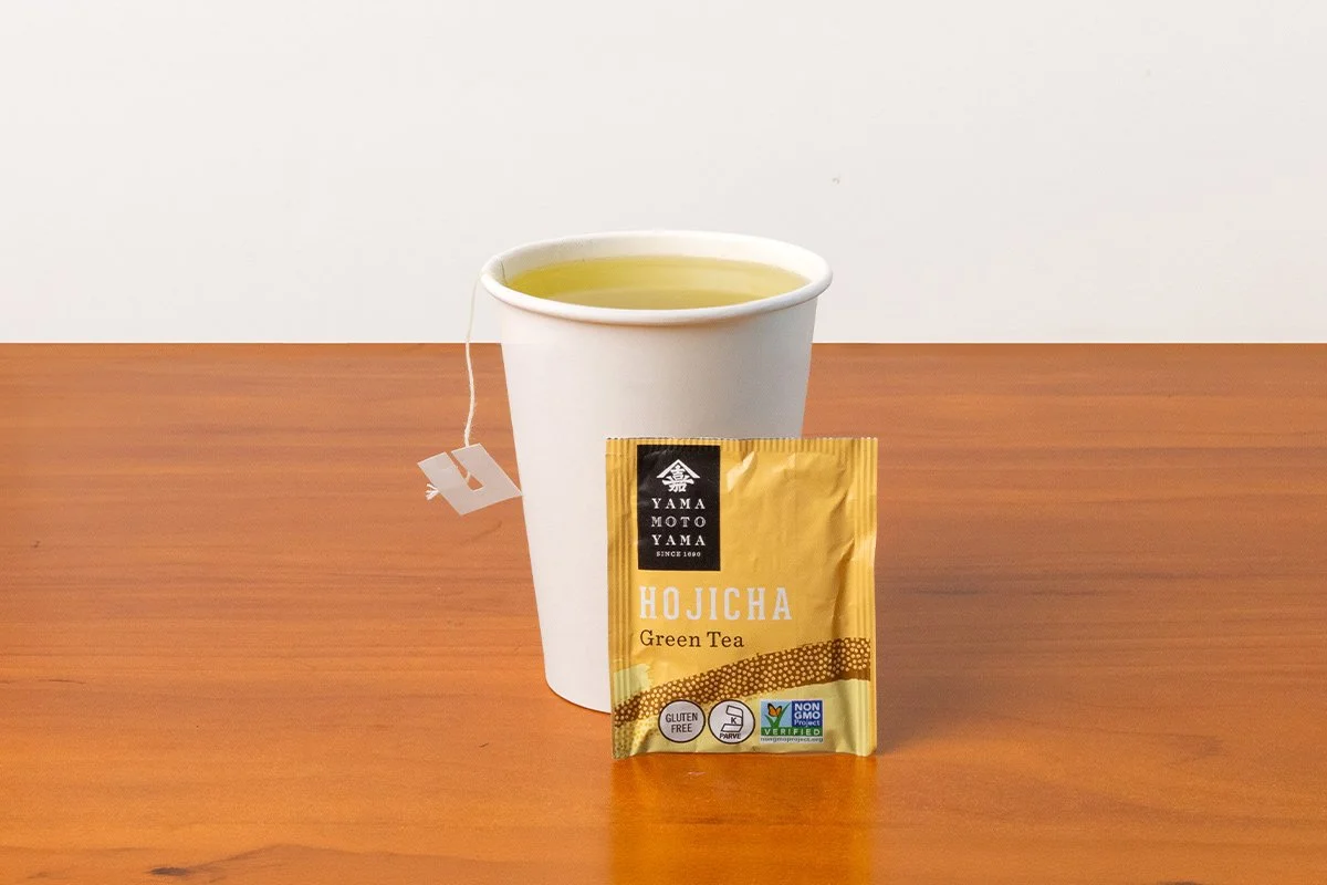 White paper cup filled with green tea and a packet of hojicha green tea placed in front of it on a wooden surface.