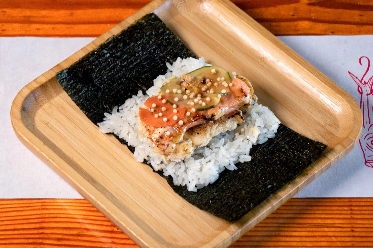 A bamboo tray showcasing an open handroll with torched crab & salmon, spicy eel sauce, pickles, and bubu arare (rice crackers) on rice (shari) and seaweed (nori).