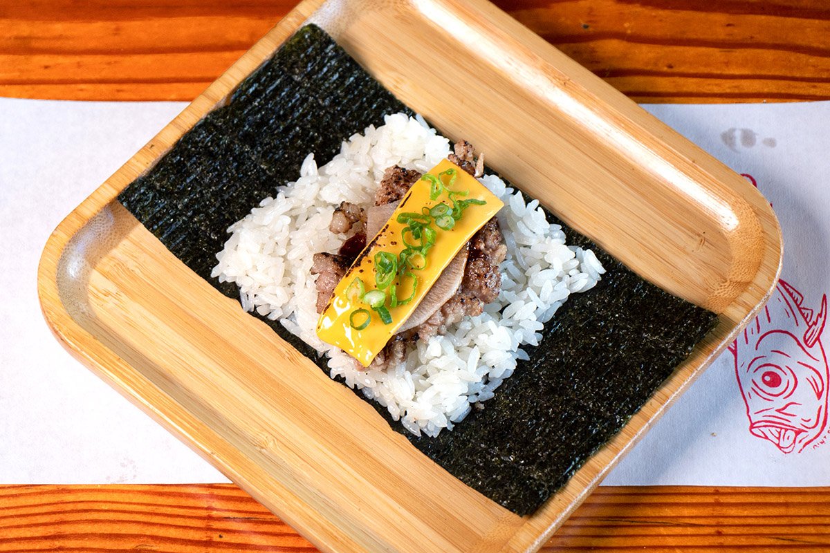 A bamboo tray showcasing an open handroll with torched a5 ground wagyu, american cheese, pickled daikon, tonkatsu sauce, scallions, on rice (shari) and seaweed (nori).