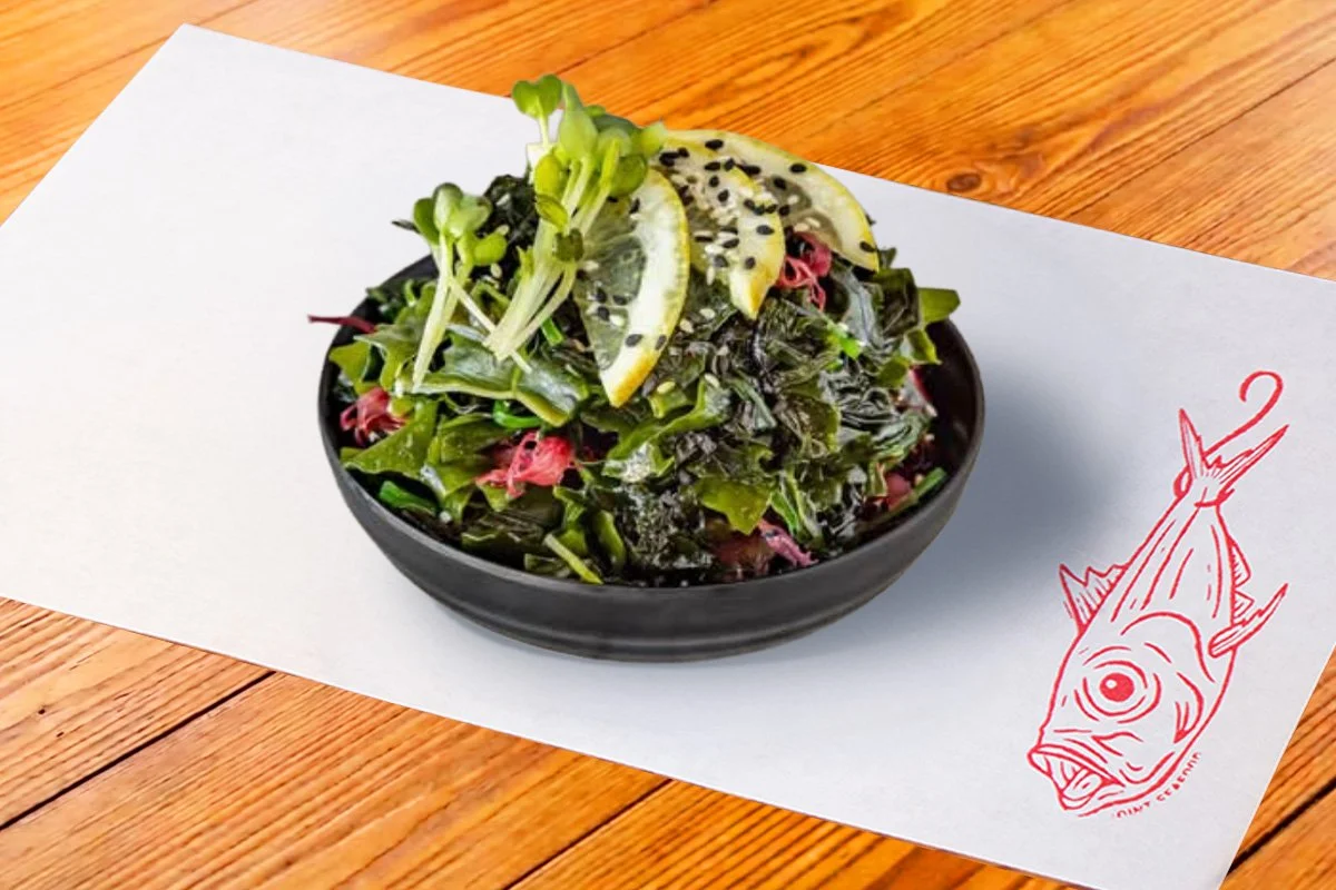 Seaweed salad topped with lemon slices, microgreens, and black sesame seeds in a black bowl on a white napkin with a red fish logo, on a wooden table.