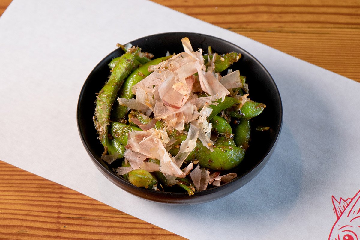 Wood table with white paper place mat with a bowl containing edamame with chili butter furikake and bonito flakes