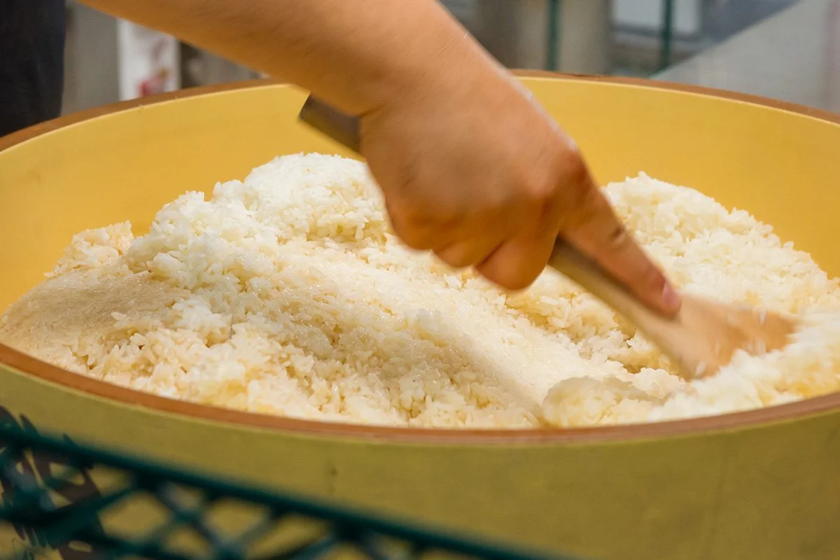 A chef preparing cooked rice seasoned with vinegar in a traditional Japanese ohitsu.