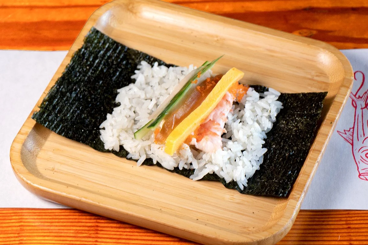 A bamboo tray showcasing an open handroll with crab, shrimp, tamago, kanpyo, cucumber, pickled daikon on rice (shari) and seaweed (nori).