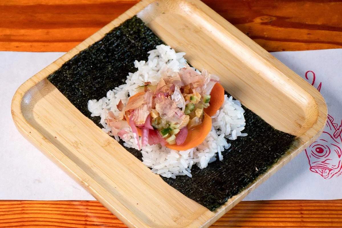 A bamboo tray showcasing an open handroll with bonito, chirashi mix, house pickles, kaiware, yukari on rice (shari) and seaweed (nori).
