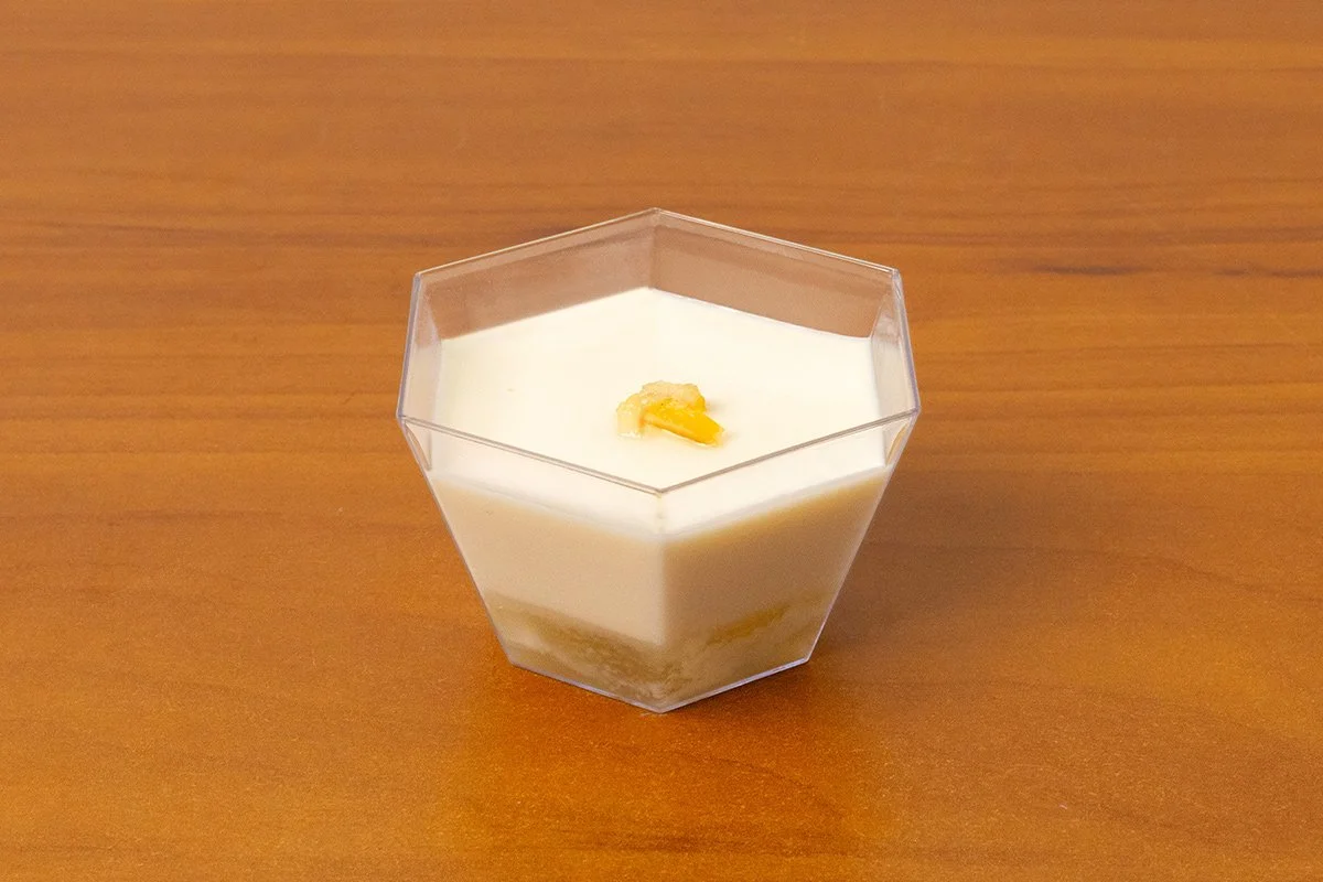 A hexagonal clear container filled with a layered yuzu panna cotta dessert, topped with a small piece of yuzu on a wooden surface.