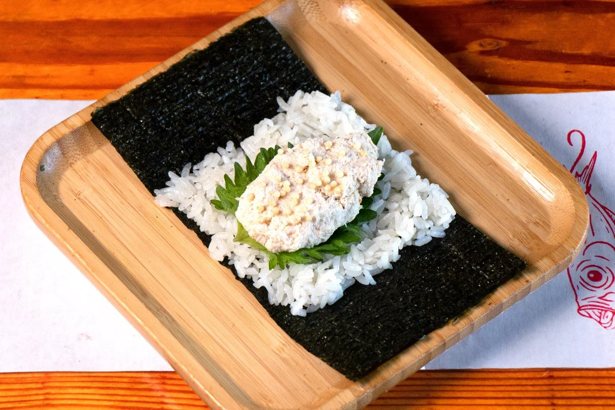 A bamboo tray showcasing an open handroll with house crab salad, shiso and bubu arare on rice (shari) and seaweed (nori).