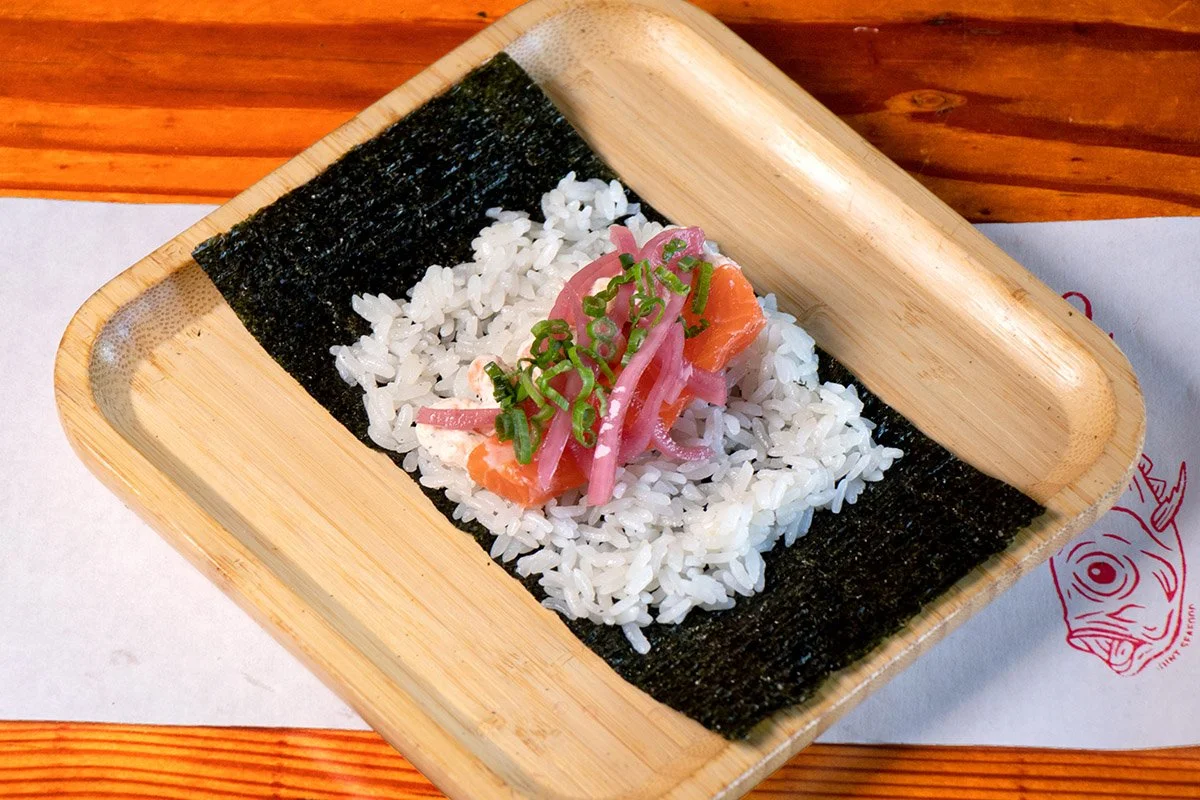 A bamboo tray showcasing an open handroll with lox shmear, steelhead, scallions, and pickled onions on rice (shari) and seaweed (nori).