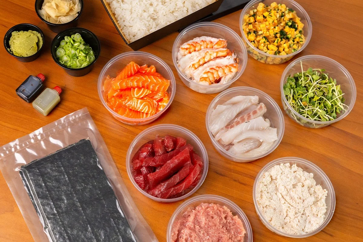 Containers of sliced raw salmon, tuna, shrimp, white fish, crab, rice, seaweed sheets, microgreens, chirashi mix, chopped green onions, wasabi, soy sauce, and pickled ginger for temaki making.