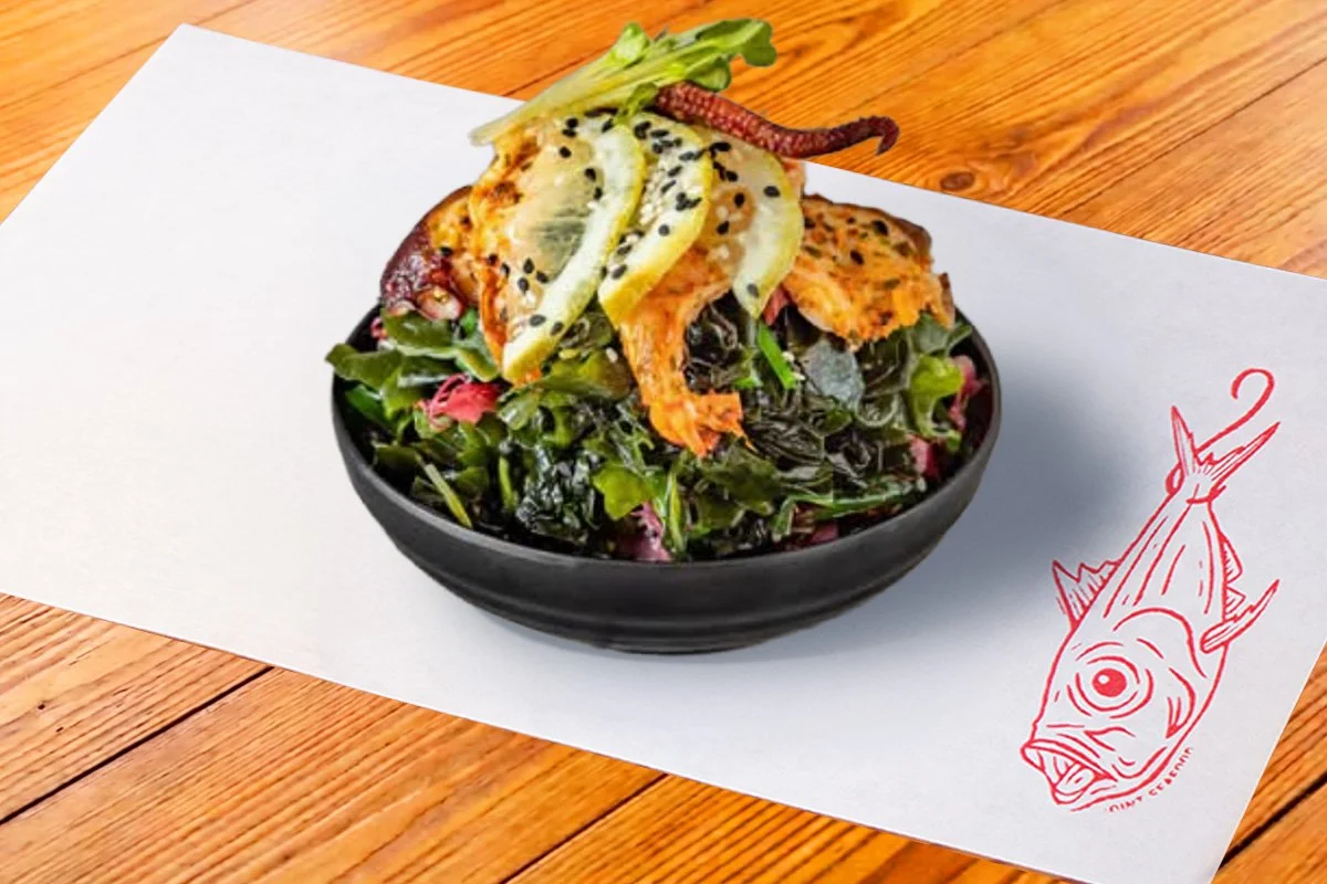Seaweed salad topped with shrimp, octopus, lemon slices, microgreens, and black sesame seeds in a black bowl on a white napkin with a red fish logo, on a wooden table.
