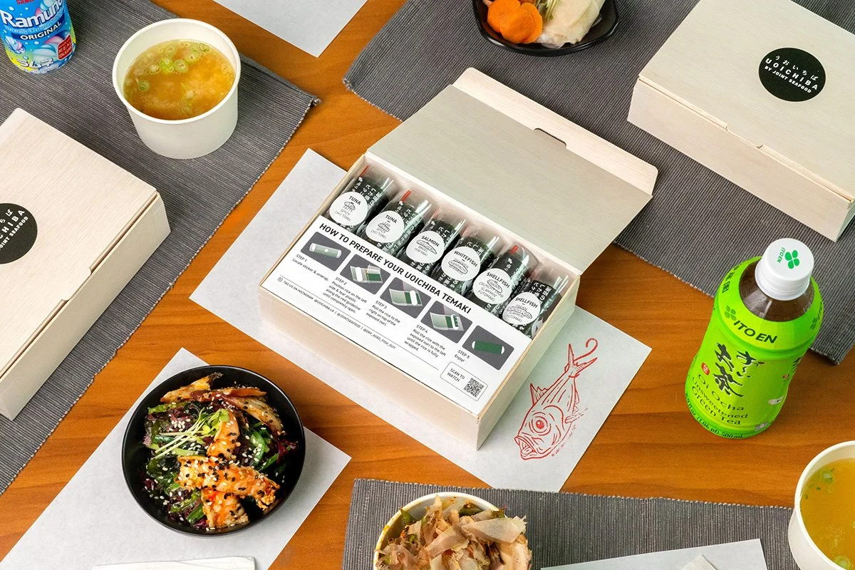 A table set with Uoichiba temaki kit, soup, seaweed salad with grilled octopus, and green tea, with the kinmedai illustration on the placemat.