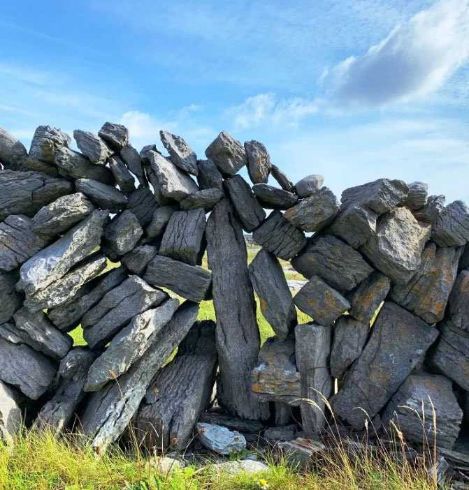 A dry stone wall constructed with irregularly shaped rocks stacked on top of each other, outside under a partly cloudy sky.