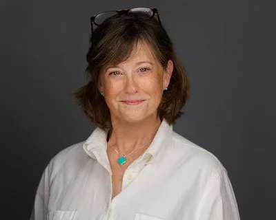 A woman with short brown hair wearing a white shirt, glasses on her head, and a turquoise necklace, smiling at the camera against a gray background.