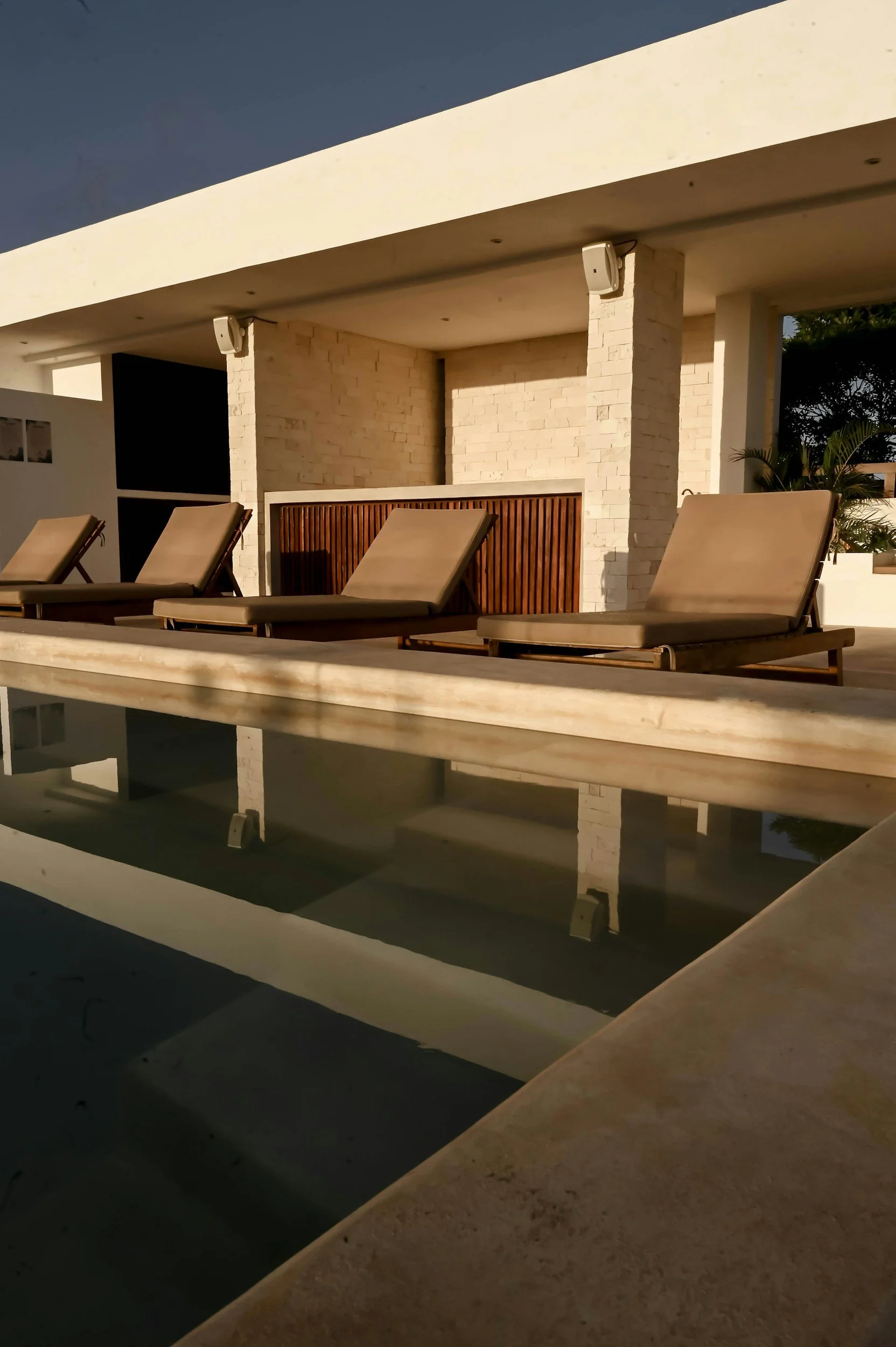 Zona de descanso en una terraza con sillones reclinables de madera y cojines beige, con paredes de ladrillo blanco y ventiladores en el techo, en una tarde soleada.