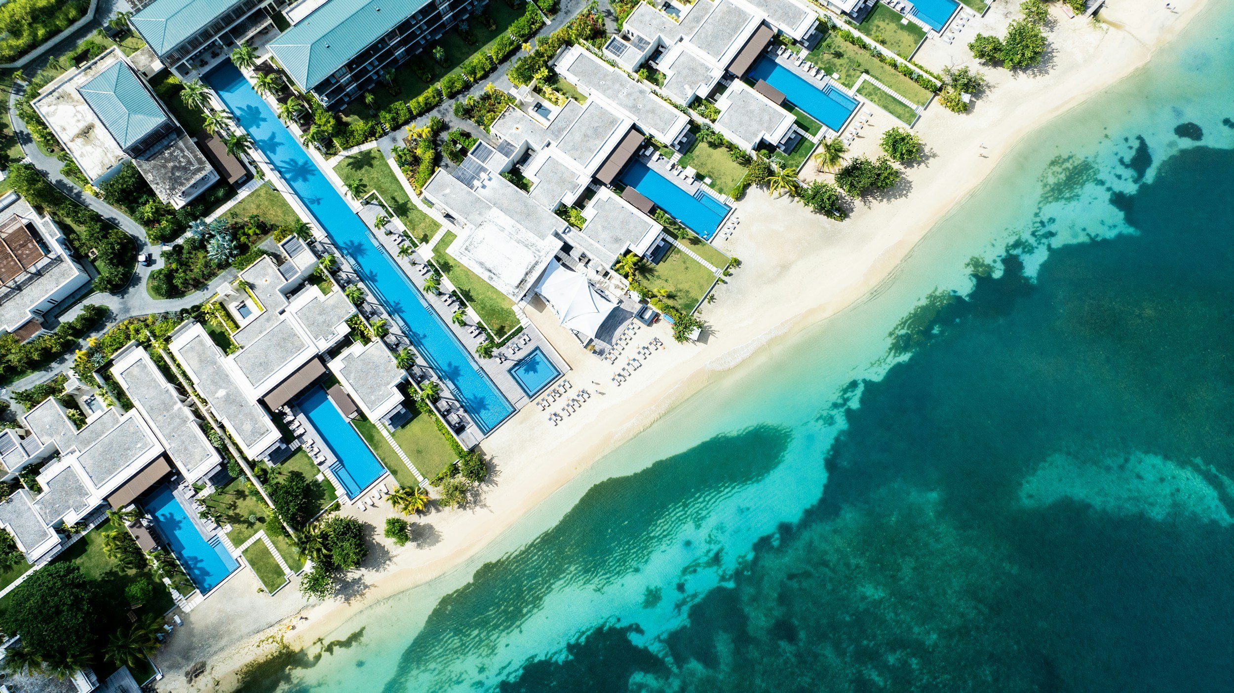 Vista aérea de un complejo turístico costero con varias piscinas, áreas verdes y edificios junto a la playa con arena y agua cristalina.