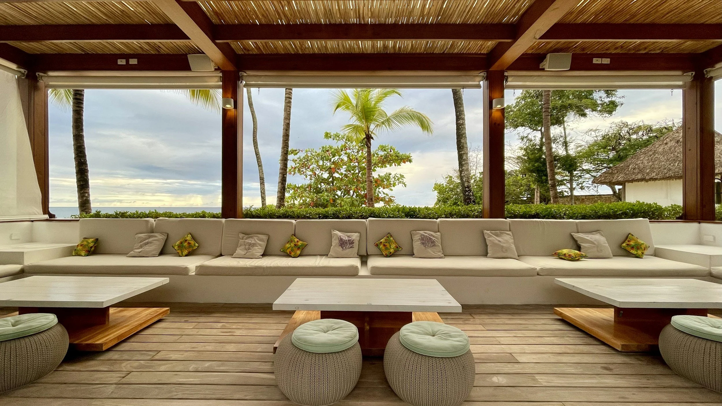 Vista desde el interior de una sala con sofá largo blanco y cojines decorativos, grandes ventanas abiertas con vista a palmeras, árboles y el mar en un entorno tropical.
