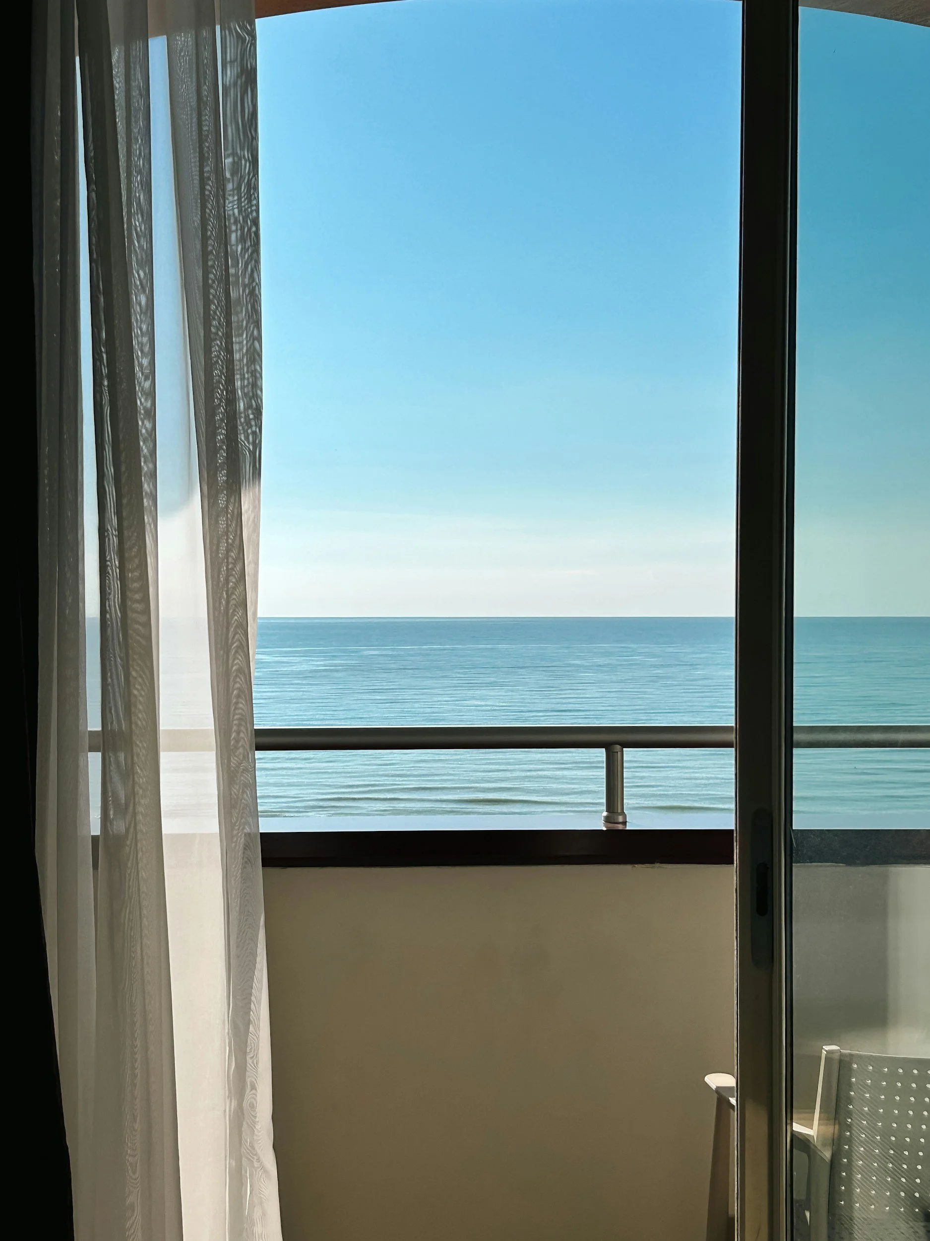 Vista del mar a través de una ventana en un cuarto, con cortinas blancas a la izquierda y una silla a la derecha.