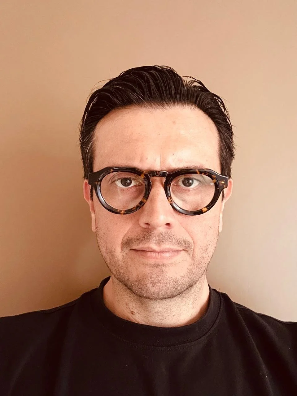 Retrato de un hombre con gafas de armazón grueso y cabello oscuro peinado hacia atrás, usando una camiseta negra, contra fondo beige.
