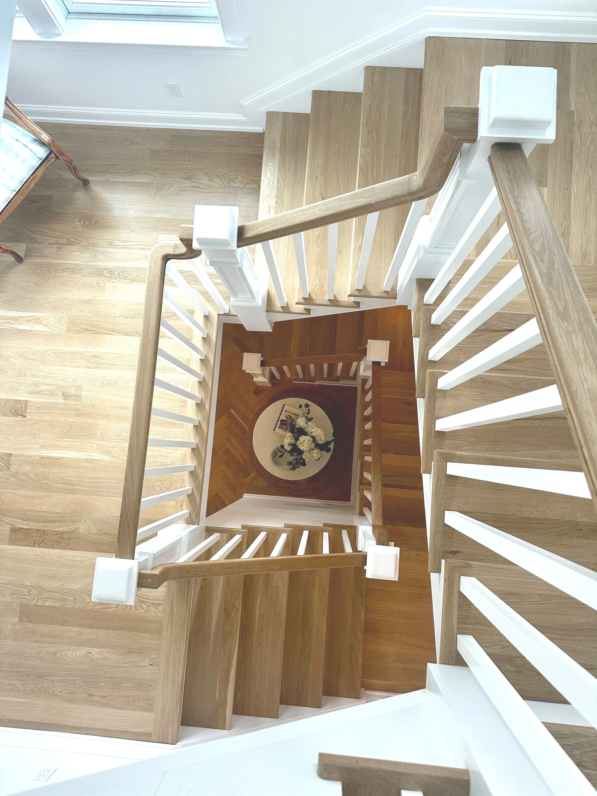 Looking down from the top of a staircase, showing wooden stairs with white railings and a landing with a round rug, flowers, and picture frames.
