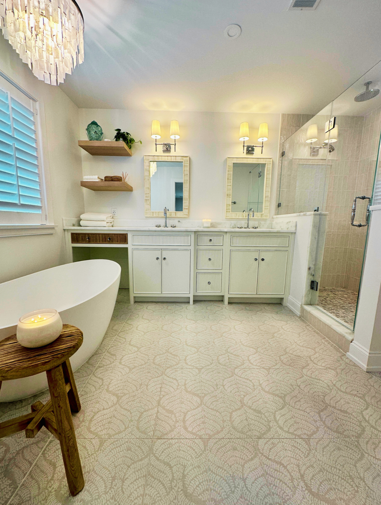 A bathroom featuring a white bathtub, a wooden stool with a lit candle, a double vanity with two mirrors and wall-mounted lights, beige tile flooring with a pattern, open shelves with towels and decorative items, and a walk-in shower with a glass doo