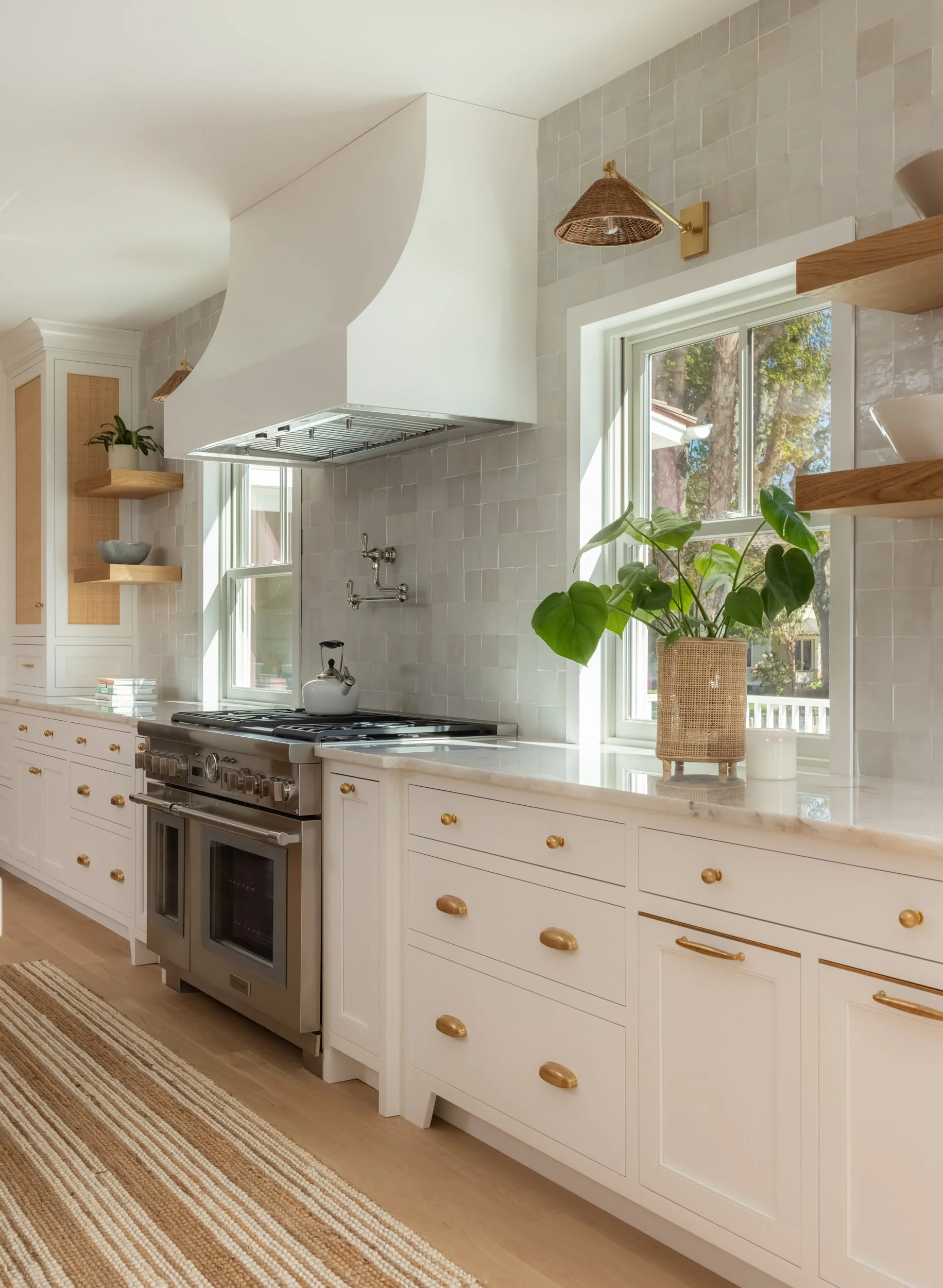 A cozy kitchen with white cabinetry, a stainless steel stove with a kettle, potted plants on open shelves, and a window with views of trees outside.