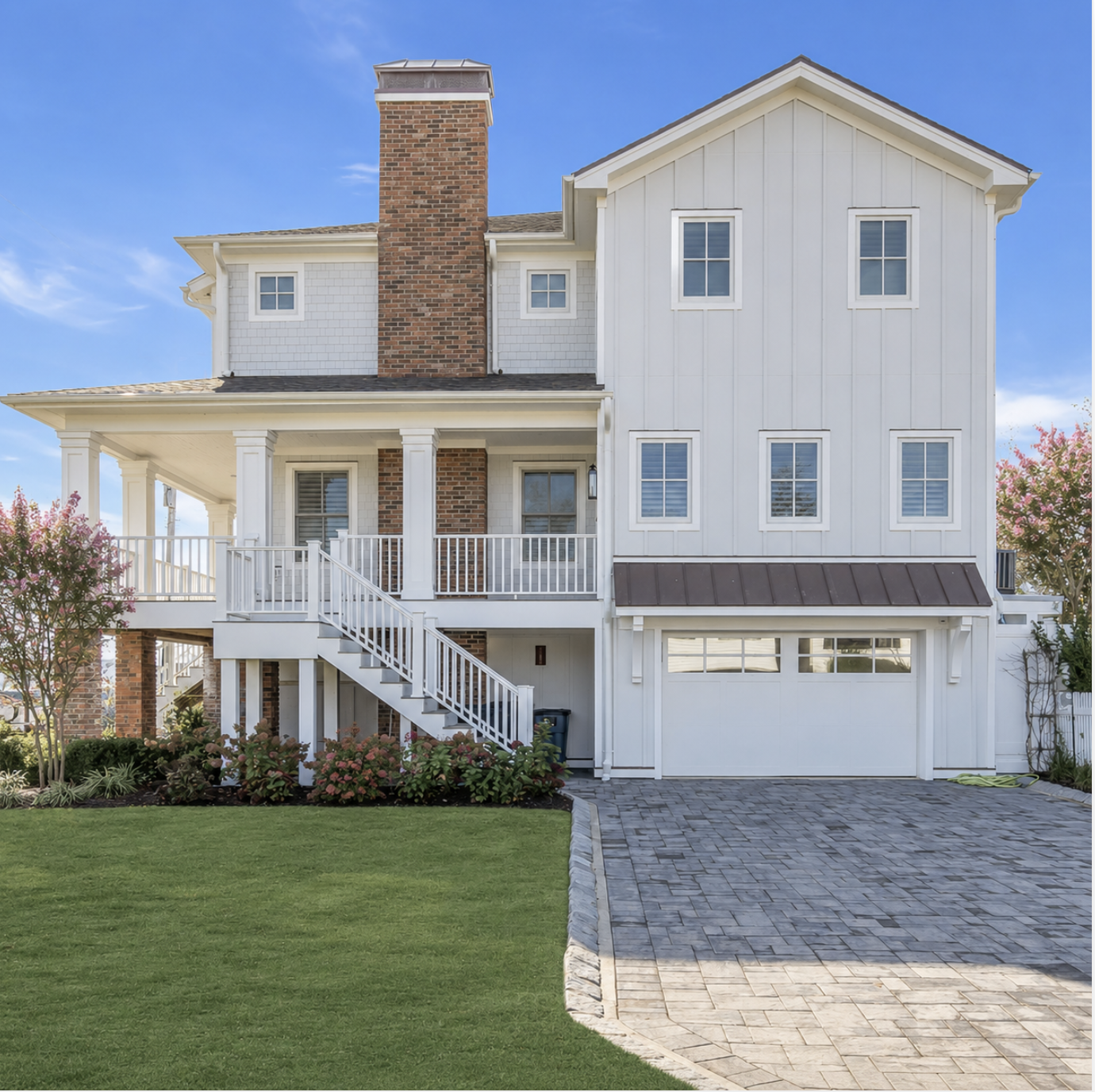 A two-story house with a white wooden exterior, a brick chimney, a front porch with stairs, and a black metal roof over the garage. The house is surrounded by grass, pink flowering trees, and a paved driveway.