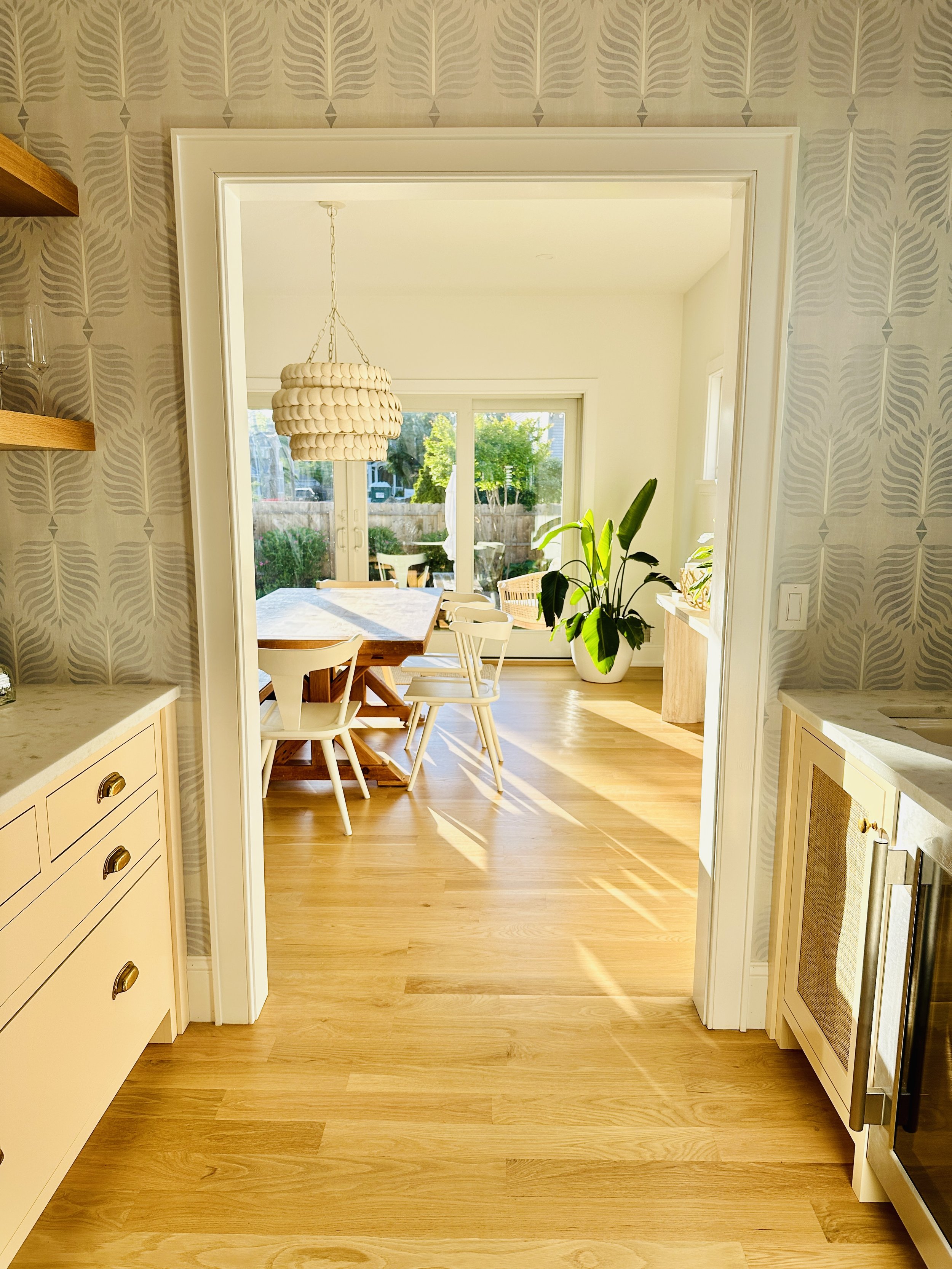 Dining area with white table and chairs, large window with view of backyard, potted plant, wood flooring, pendant light above table, wallpapered walls.