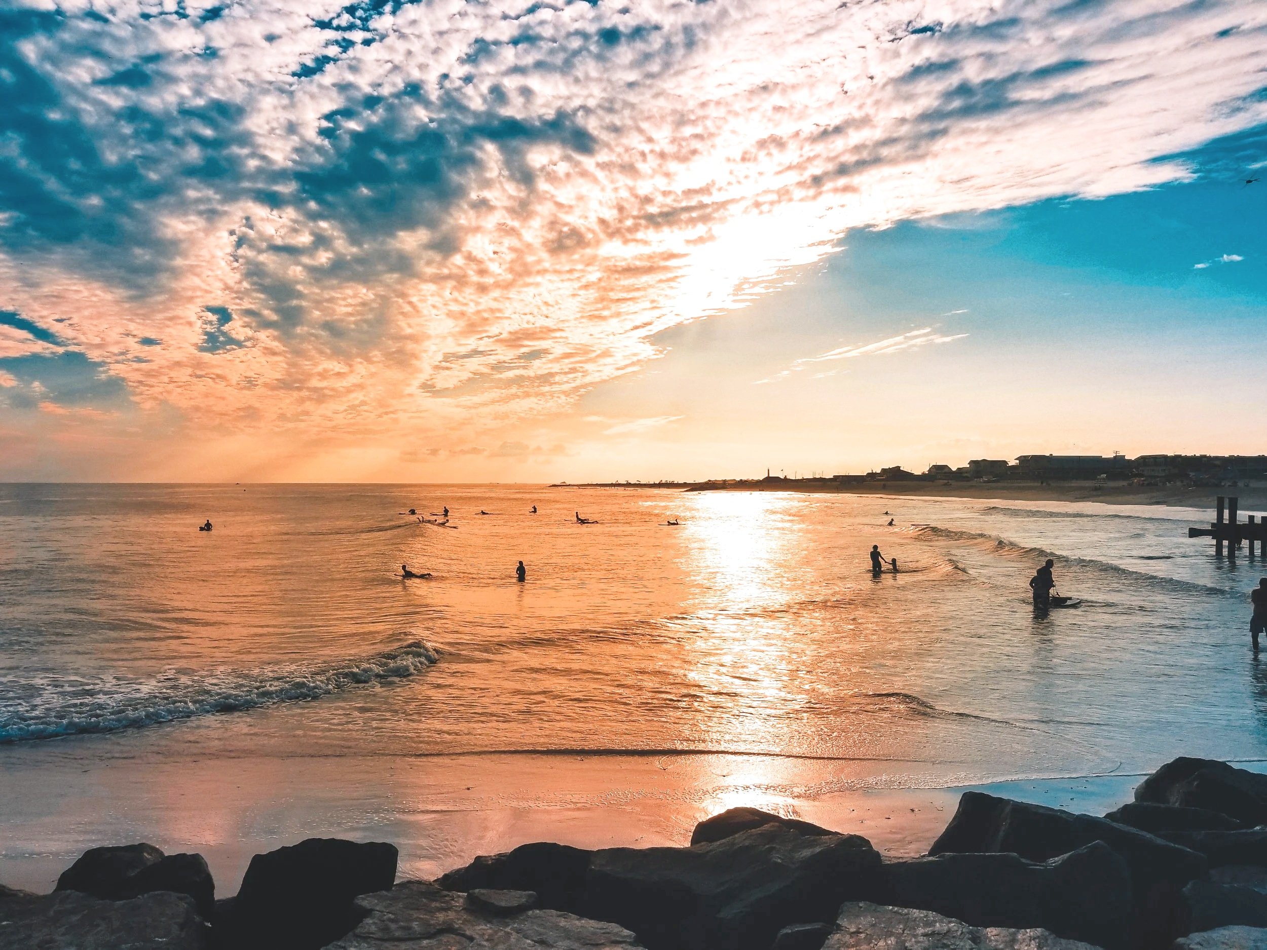 A beach scene at sunset with a sky filled with colorful clouds, surfers in the water, and rocks along the shoreline.