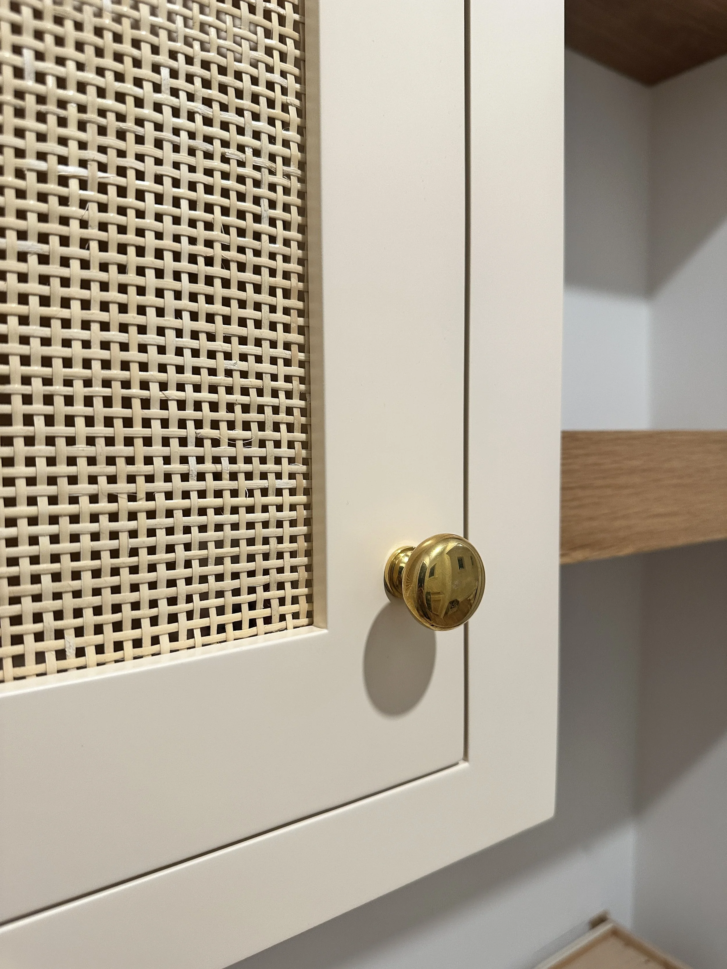 Close-up of a white cabinet with a woven rattan door and a brass doorknob, situated against a wall with a wooden shelf visible.