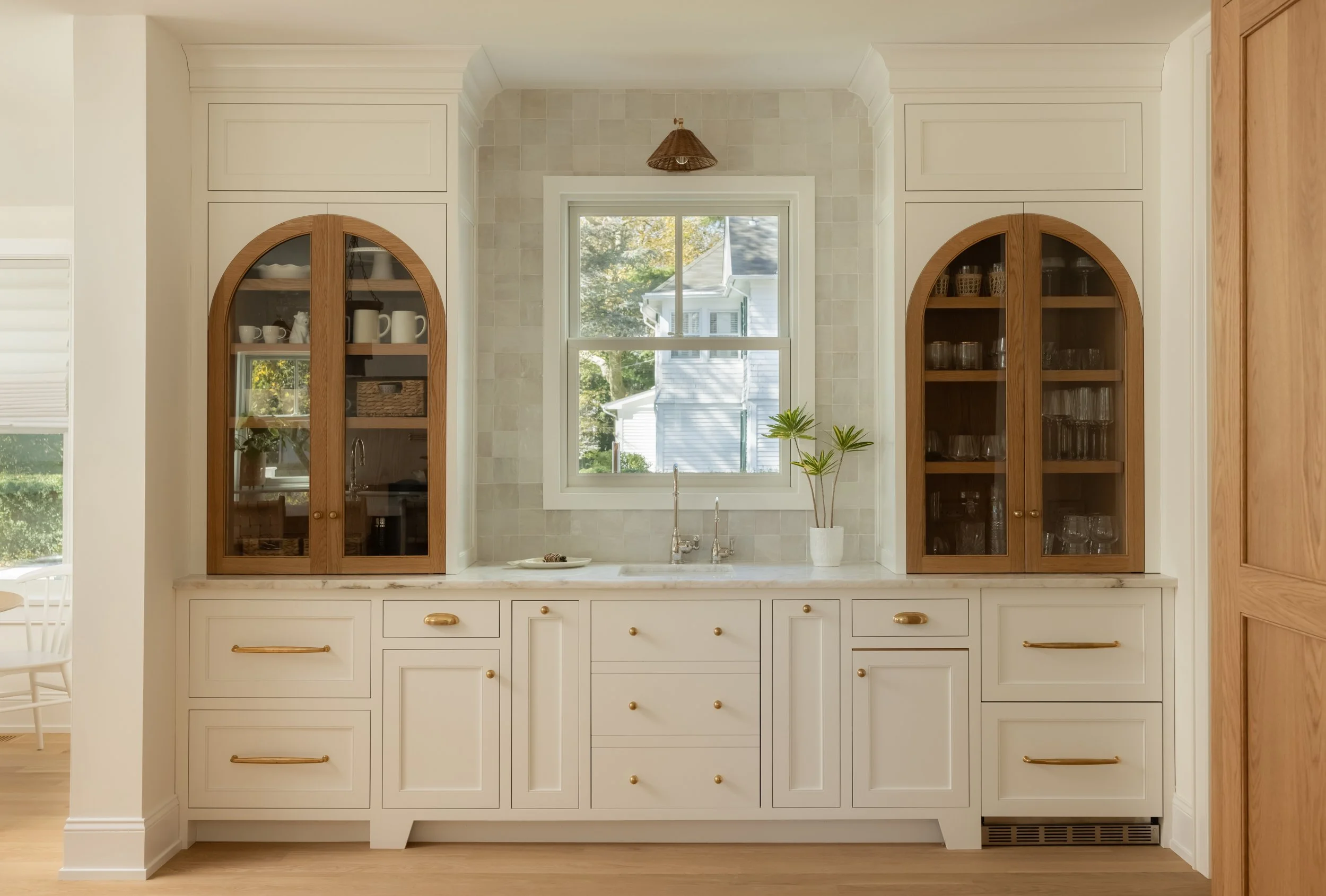 Kitchen with white cabinets, wooden glass-paneled doors, a window with a view of trees and a house, a small potted plant, and a marble countertop.