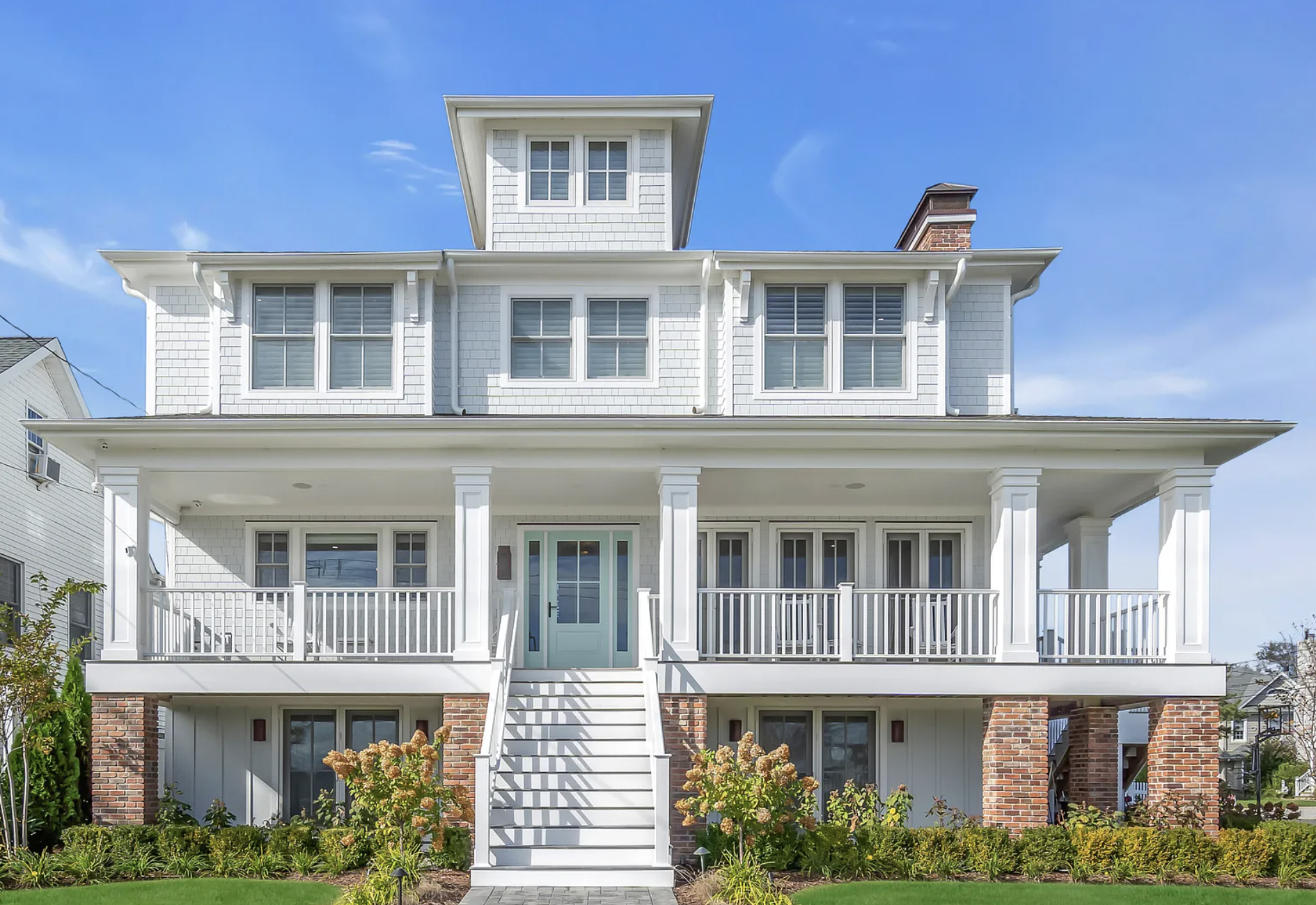 Front view of a large, white, three-story house with a porch, stairs, and multiple windows, with a landscaped garden and clear blue sky.