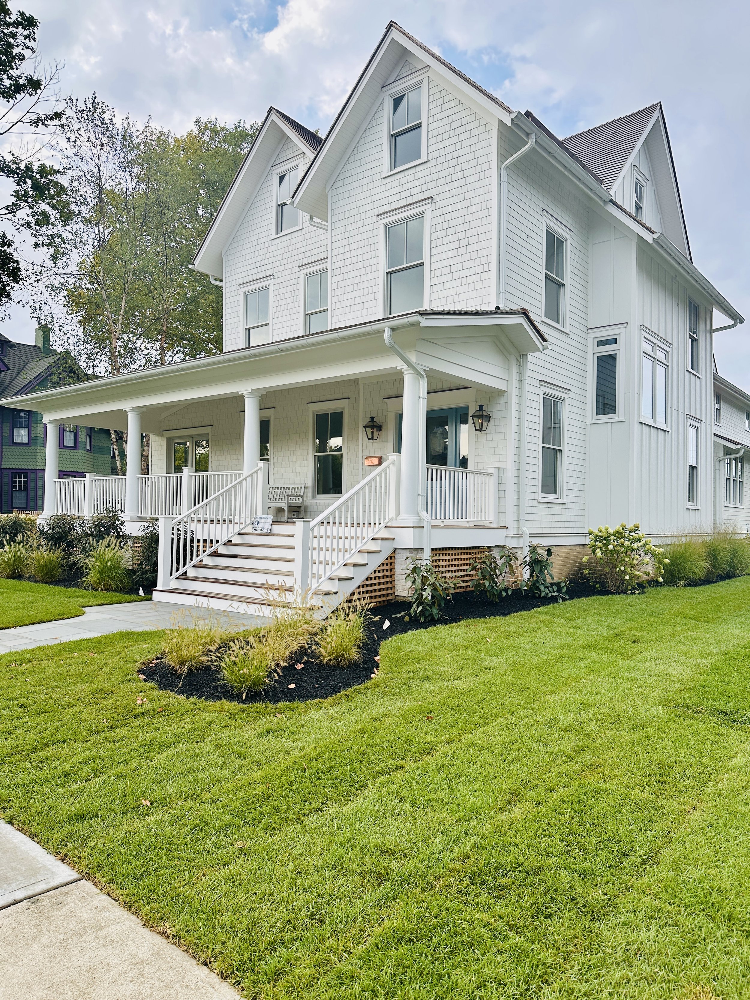 A white, multi-story house with a front porch, porch lanterns, and stairs, surrounded by a well-maintained lawn, flower beds, and a partly cloudy sky.
