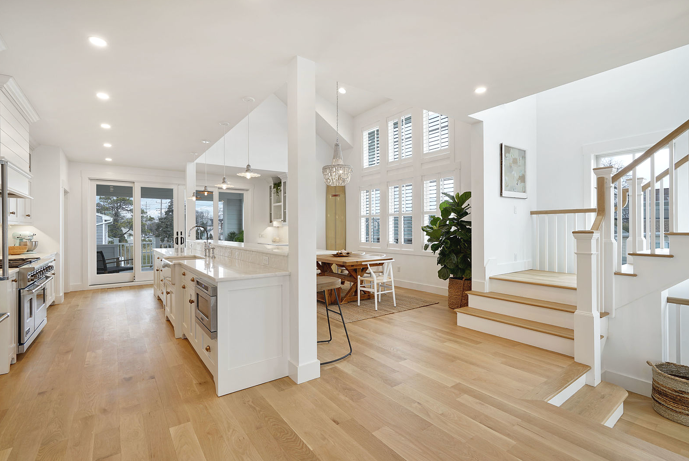 Bright, open-concept kitchen and dining area with white cabinetry, a large island, wooden flooring, and large windows with shutters, adjacent to staircase with white balusters.